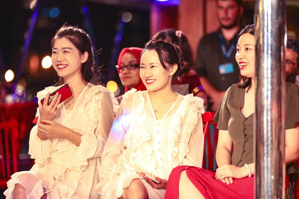 한 행사에 세 명의 여성이 앉아 미소를 짓고 있습니다. 하나는 빨간색 전화기를 들고 있습니다. 다른 참석자는 백그라운드에서 볼 수 있습니다.