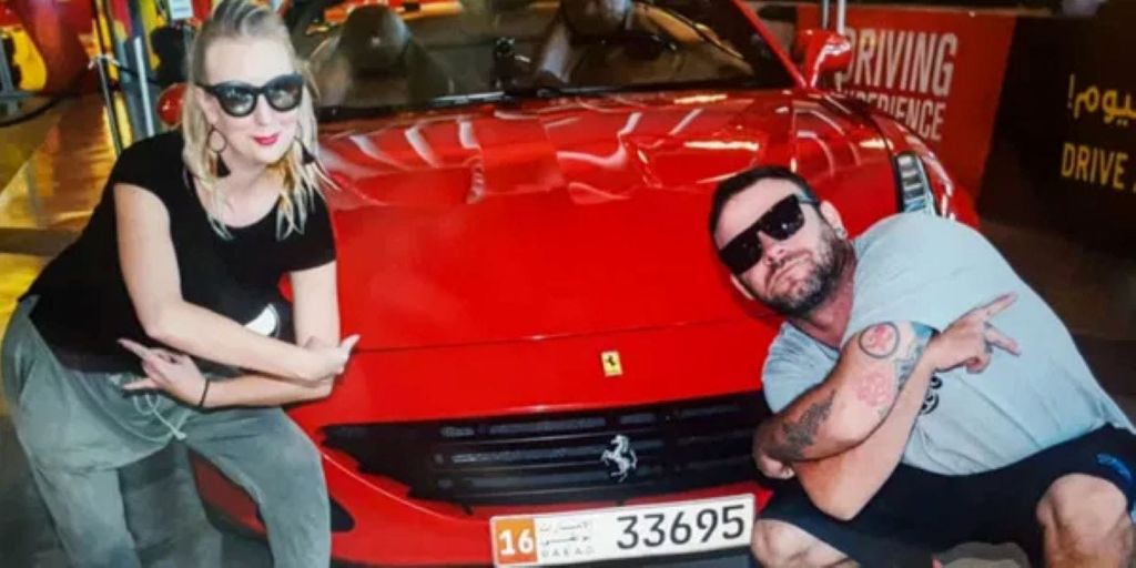 선글라스와 캐주얼한 옷을 입은 여성이 앞판에 숫자 16이 새겨진 빨간색 스포츠카 옆에서 포즈를 취하고 있습니다.