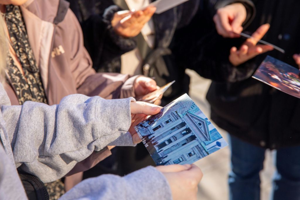 사람들은 인쇄된 사진과 문서를 들고 있는데, 하나는 기둥과 삼각형 페디먼트가 있는 건물을 보여줍니다.