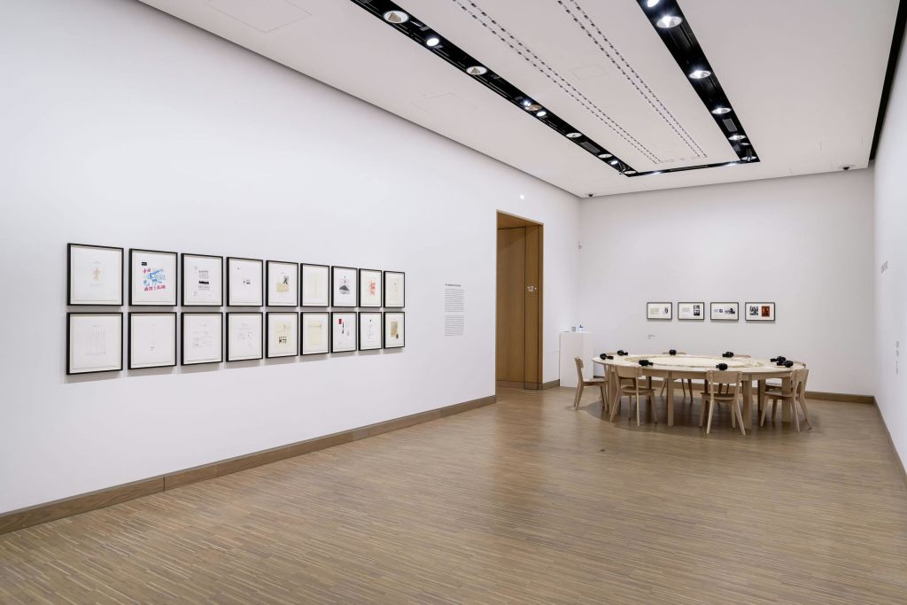 흰색 벽에 액자가 놓인 미술품, 나무 바닥, 구석에 의자가 놓인 테이블이 있는 현대적인 갤러리 객실입니다.