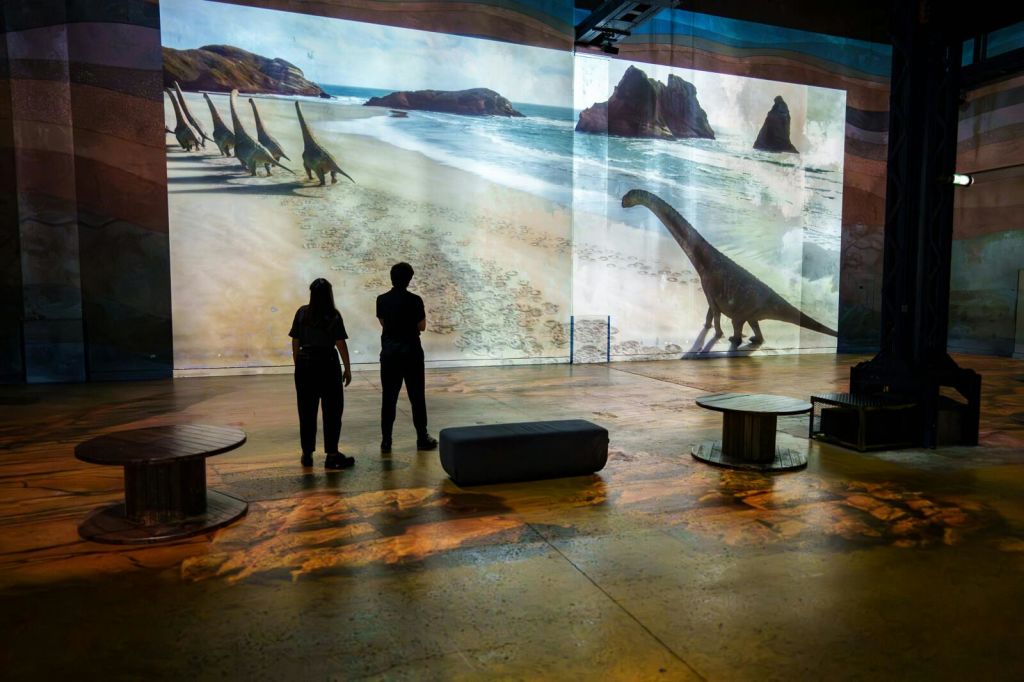 두 사람이 갤러리에 서서 해변을 거닐고 있는 공룡들의 커다란 투영을 관찰하고 있으며, 주변에는 좌석과 인테리어 소품이 놓여 있습니다.
