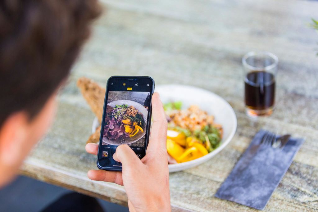 스마트폰을 들고 도금한 식사를 나무 테이블에 올려놓고 사진을 찍는 사람. 진한 음료가 담긴 잔이 근처에 있습니다.