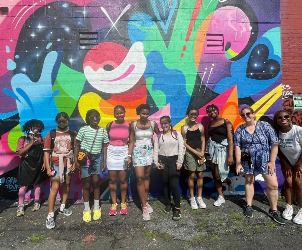 한 무리의 사람들이 화창한 날 다채로운 색상의 그래피티로 덮인 벽 앞에 서서 미소를 지으며 사진을 찍으며 포즈를 취하고 있습니다.