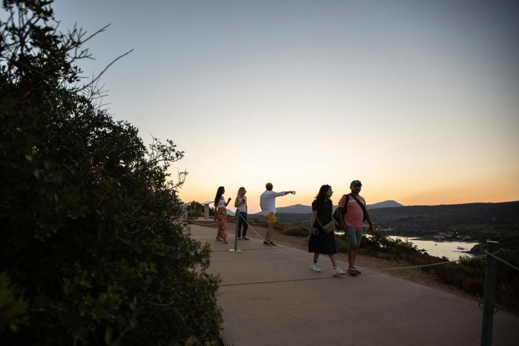 수니오곶에서 일몰을 향해 걷는 관광객들. 퇴색한 하늘 아래 낮은 수풀 옆 길을 따라 산책하는 사람들.