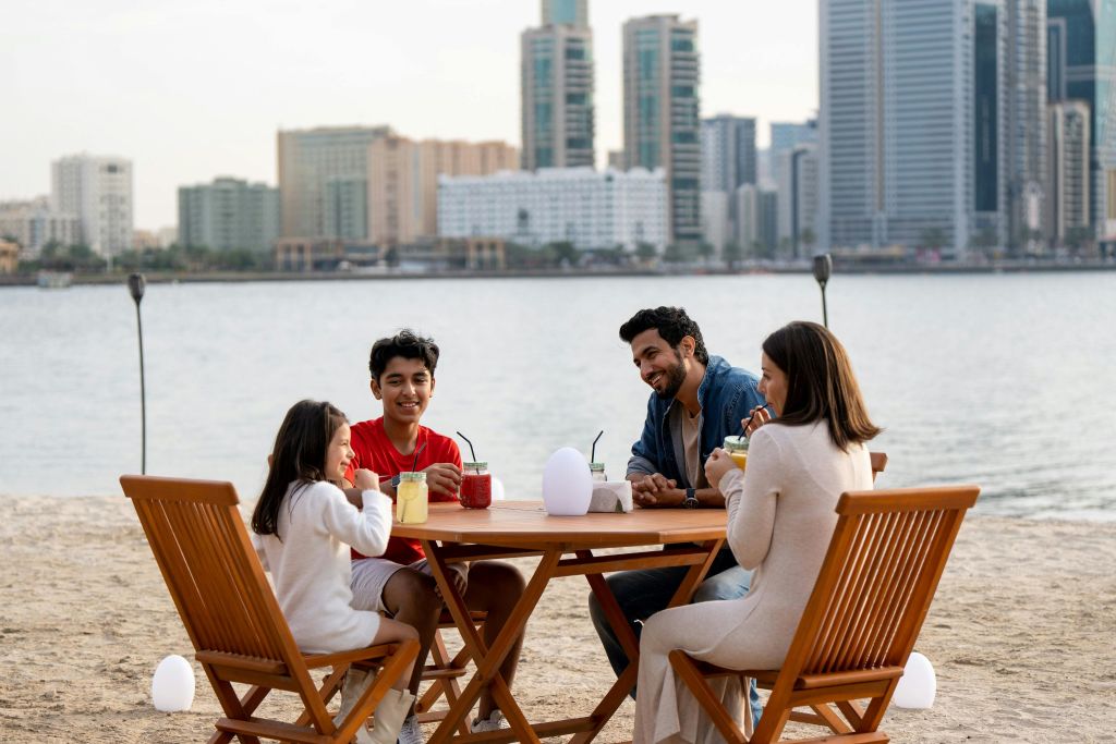 4명이 해변의 나무 테이블에 앉아 도시 풍경과 바다를 배경으로 앉아 있습니다. 음료와 음식이 테이블에 있습니다.