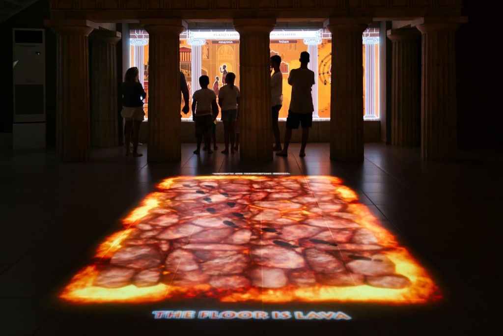 전시회에서 "The Floor is Lava" 를 묘사한 인터랙티브 디스플레이를 관찰하는 사람들로, 생동감 넘치는 배경에 실루엣이 보입니다.