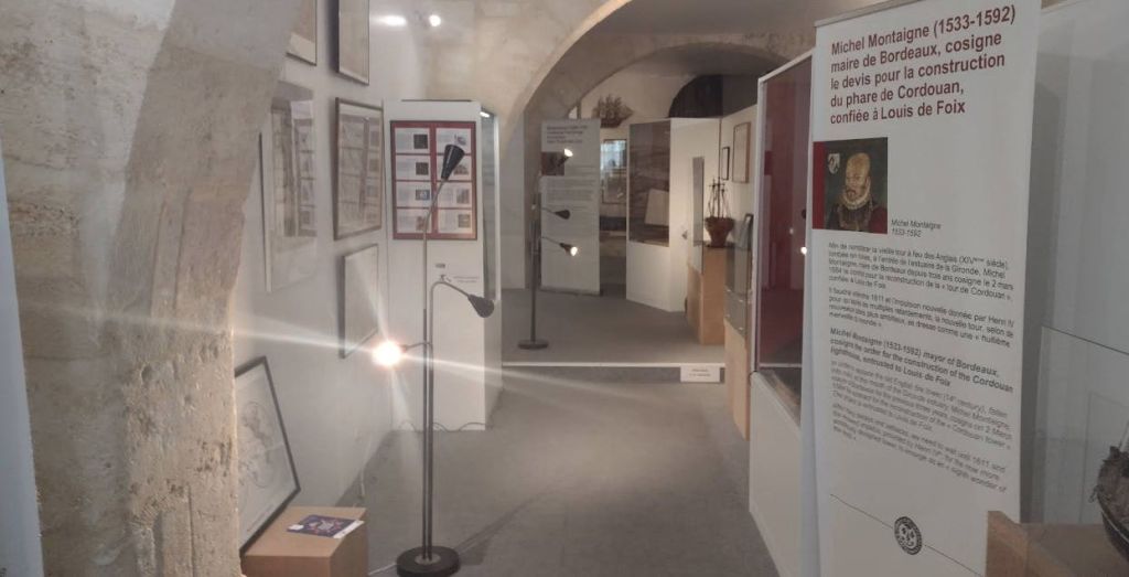 밝은 공간에 벽, 정보 패널, 전시 케이스 및 플로어 램프에 액자 예술 작품을 전시하는 박물관 전시관입니다.