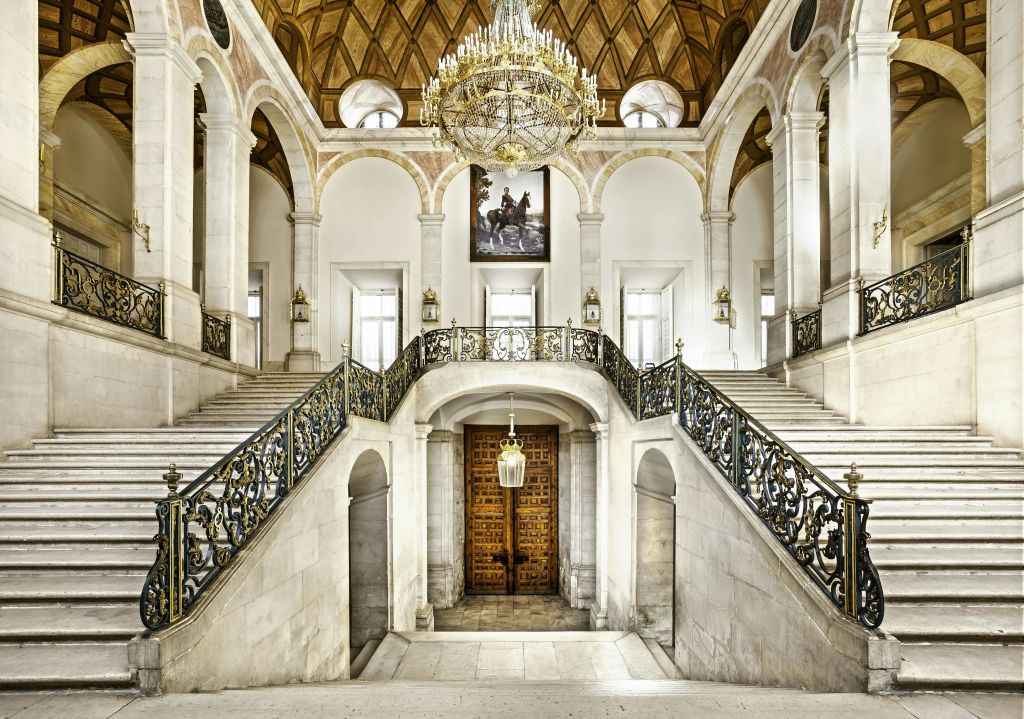 화려한 난간이 있는 웅장한 대리석 계단, 커다란 샹들리에, 아치형 창문, 그림 아래 나무 문이 있습니다.