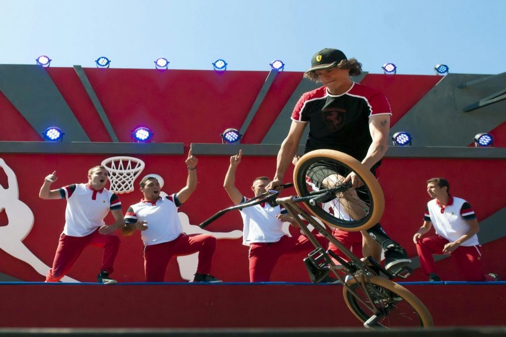 무대에서 BMX 라이더가 스턴트를 선보이고, 빨간 바지와 흰색 셔츠를 입은 댄서 4명이 무대에서 환호합니다.