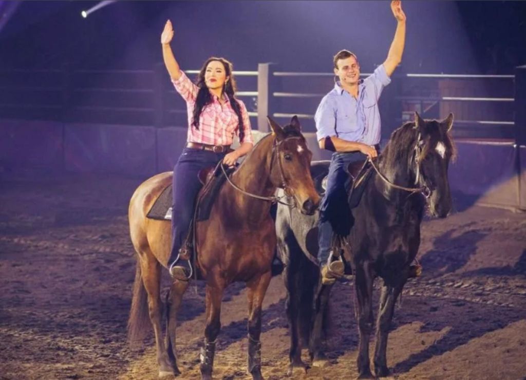 경기장에서 말을 타고 관객에게 손을 흔드는 두 사람. 여자는 분홍색 격자무늬 셔츠를 입고, 남자는 파란색 셔츠를 입습니다.