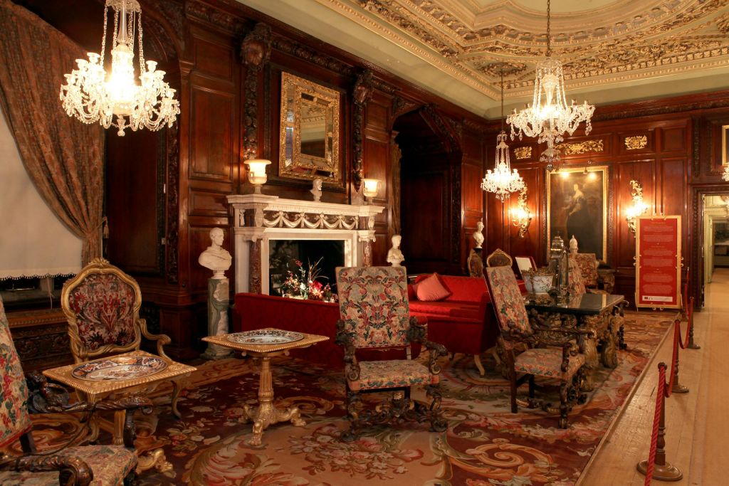 장식된 벽난로, 빈티지 가구, 샹들리에, 조각상이 있는 화려한 객실입니다. 디테일한 목재 패널과 무늬가 있는 카펫.