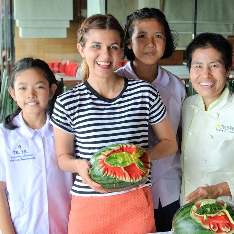 네 명이 서서 웃고 있으며, 한 사람이 새겨진 수박을 들고 있습니다. 테이블에는 더 많은 새겨진 수박이 놓여 있습니다.