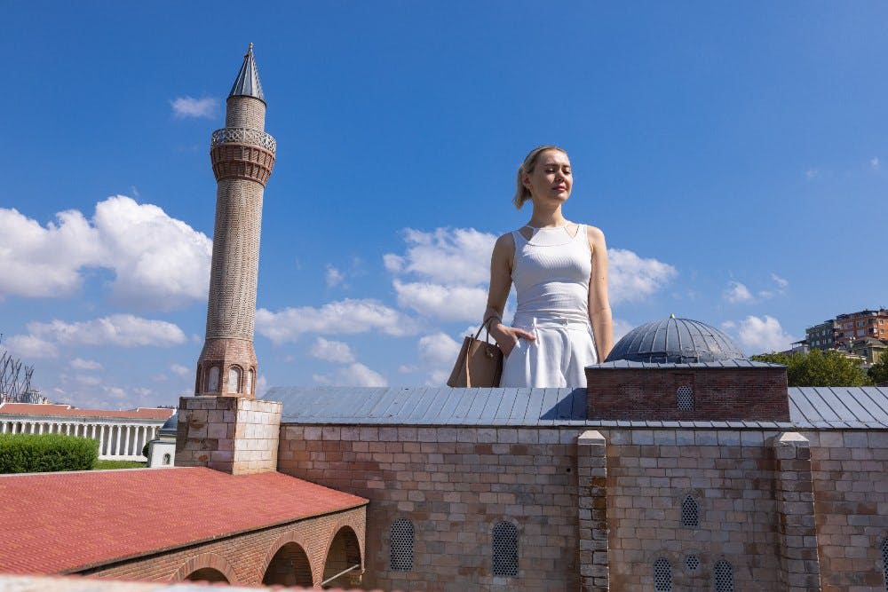 흰색 드레스를 입은 한 여성이 맑고 푸른 하늘 아래 돔이 있는 벽돌 건물 모형 뒤에 서 있습니다.