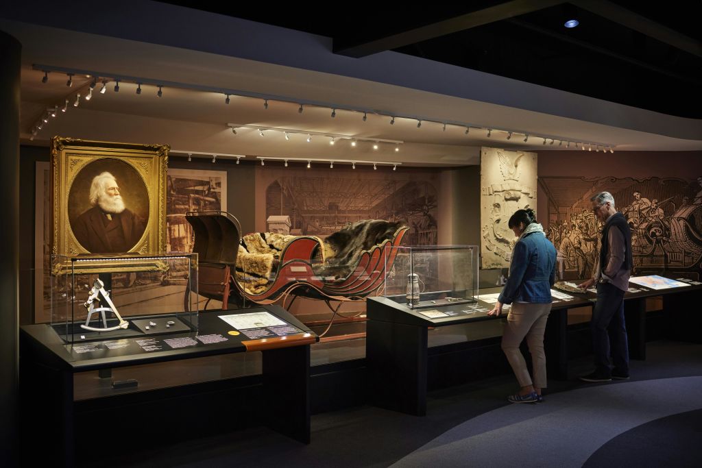 역사적인 썰매, 유물, 부드러운 조명 아래 액자에 담긴 초상화를 포함한 박물관 전시를 관람하는 사람.