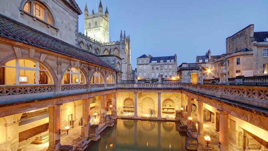 아치형 구조물로 둘러싸인 중앙 수영장이 있는 유서 깊은 로마식 목욕탕으로, 높은 교회 타워가 배경으로 보입니다.