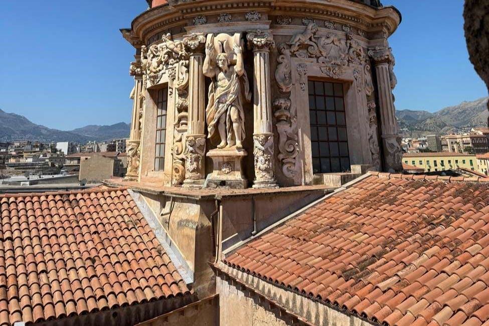 붉은 기와 지붕과 함께 동상과 기둥이 있는 화려한 석조 건물. 배경에는 먼 산과 하늘이 포함됩니다.