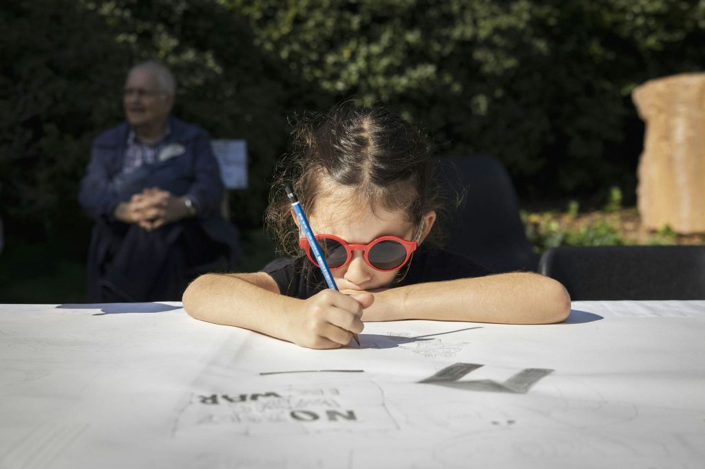 야외에서 빨간색 선글라스를 쓴 어린 소녀가 커다란 종이에 그림을 그리고 있으며, 배경에는 나이든 남성이 앉아 있습니다.