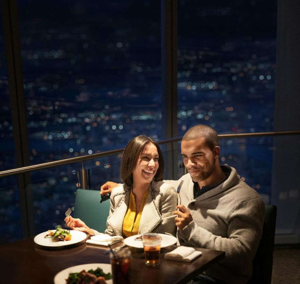 밤에 커다란 창문으로 도시 스카이라인이 보이는 레스토랑의 테이블에서 한 남성과 한 여성이 함께 식사를 하고 있습니다.