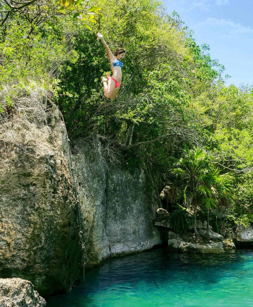 수영복을 입은 사람이 바위 절벽에서 뛰어내려 무성한 녹음으로 둘러싸인 맑고 푸른 물 속으로 들어갑니다.
