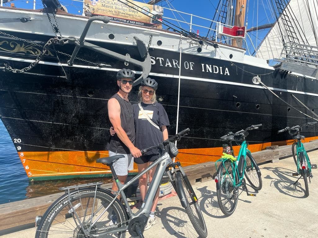 화창한 날 항구에 정박한 "인도의 별" 배 앞에 헬멧을 쓴 두 사람이 자전거를 타고 서 있습니다.