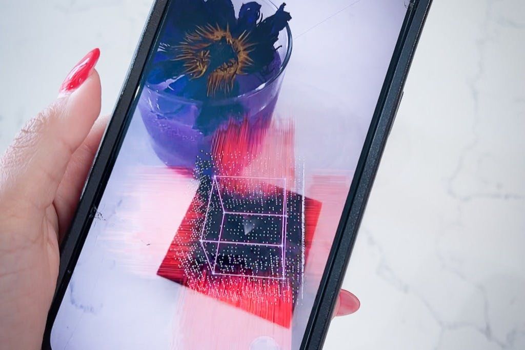 보라색 음료를 꽃과 겹쳐놓은 큐브 그래픽의 증강 현실 화면을 보여주는 스마트폰 화면입니다.