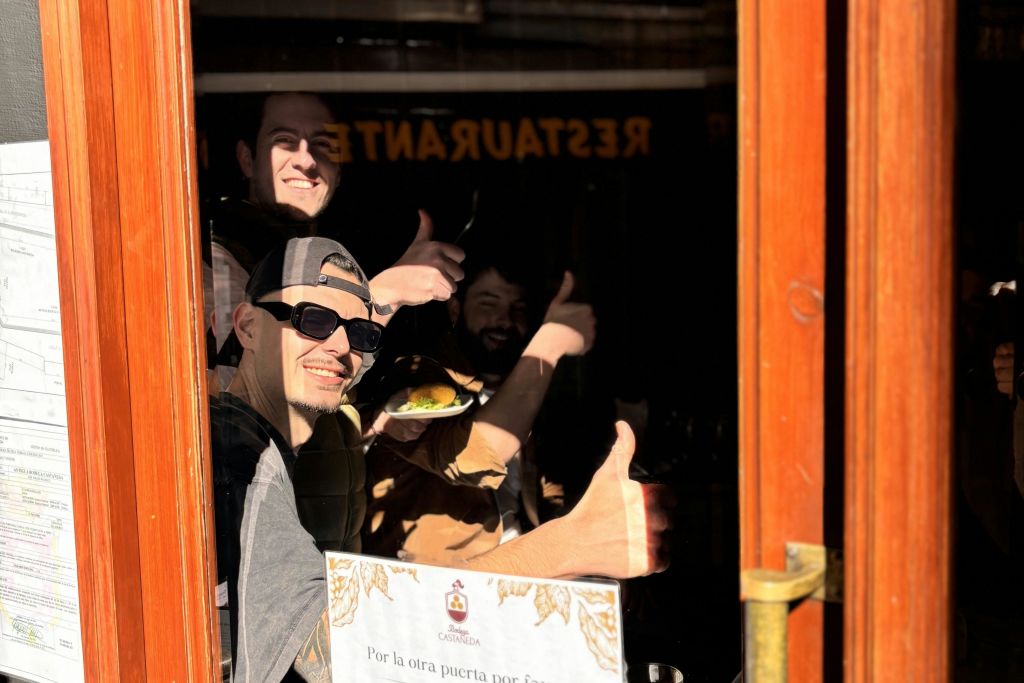 레스토랑 창문 뒤에서 세 사람이 엄지손가락을 치켜세우고 한 사람은 접시를 들고 있습니다. 창에 표시된 메뉴와 표지판.