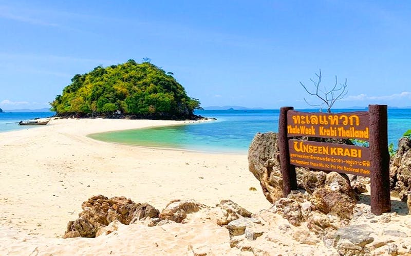 맑은 물, 모래 해변, 바위 전경, 라틴어가 아닌 문자로 된 표지판이 있는 해변. 배경에는 섬이 보입니다.