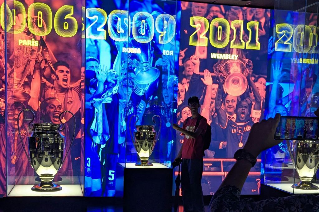 박물관에 세 개의 UEFA 챔피언스 리그 트로피가 전시되어 있으며, 다채로운 배경과 한 사람이 한 곳을 가리키고 있습니다.