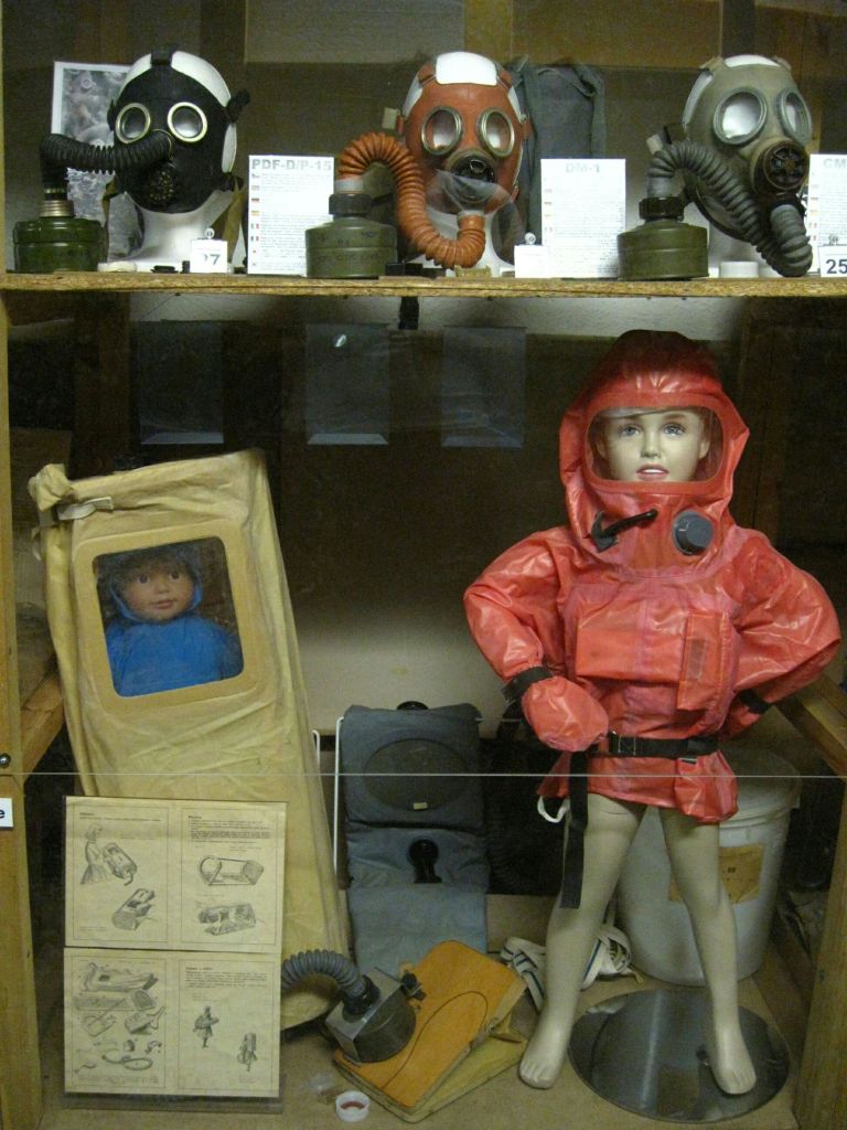 방독면과 보호 장비가 달린 마네킹, 플라스틱으로 감싸인 파란색 수트를 입은 어린이 마네킹이 전시되어 있습니다.