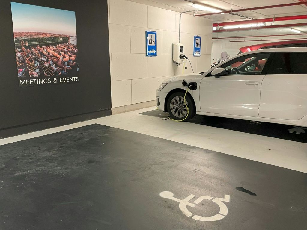 주차장의 전기차 정류장에서 흰색 차량이 충전 중입니다. 근처 공간에는 휠체어 기호가 표시되어 있습니다.