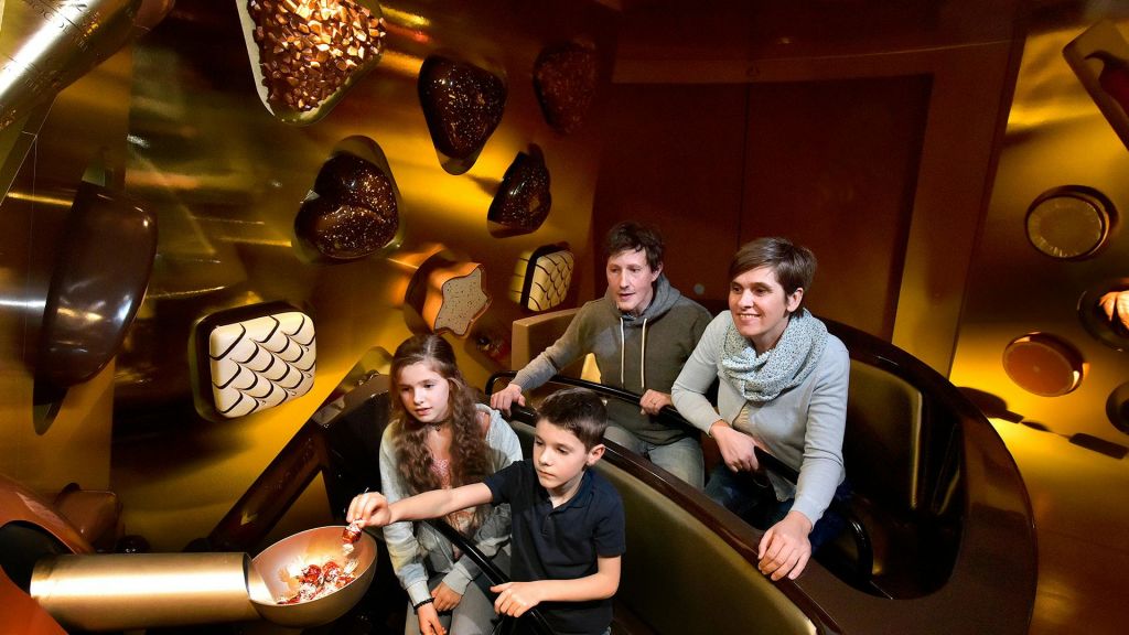 커다란 하트 모양과 다른 초콜릿 틀로 장식된 벽이 있는 어두운 어트랙션에 4인 가족이 있습니다.