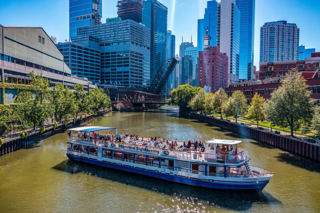 "해안선 관광" 표지판이 있는 투어 보트는 높은 도시 건물과 높이 솟은 도개교로 둘러싸인 강을 따라 이동합니다.