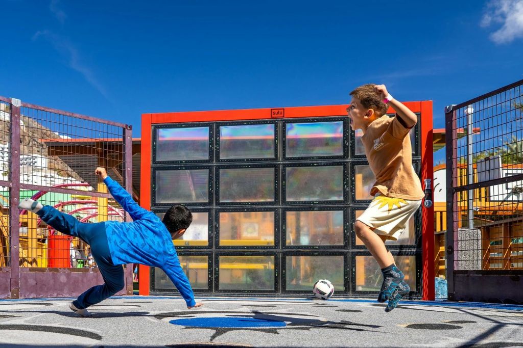 두 명의 어린이가 현대적이고 투명한 패널 벽과 밝은 주변 환경을 갖춘 다채로운 야외 코트에서 축구를 하고 있습니다.