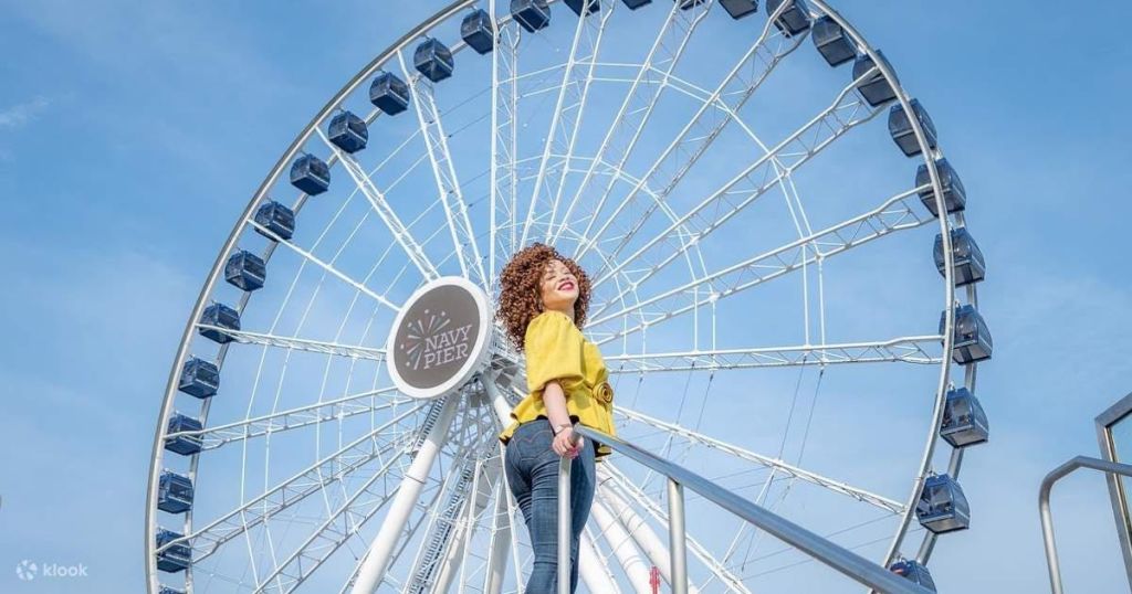 화창한 날 노란색 재킷과 청바지를 입은 미소 짓는 여성이 네이비 피어 대관람차 앞에 서 있습니다.