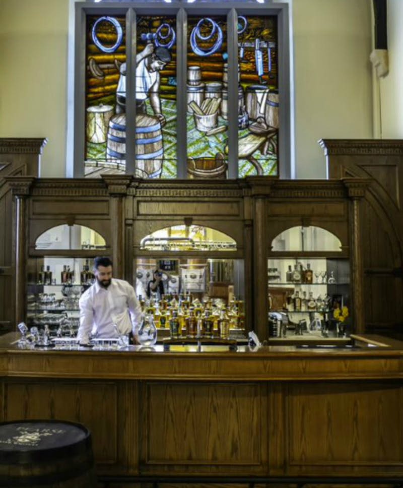 와인 통과 포도가 그려진 스테인드글라스 창문 아래 다양한 병과 잔이 놓인 나무 바 뒤에 바텐더가 서 있습니다.