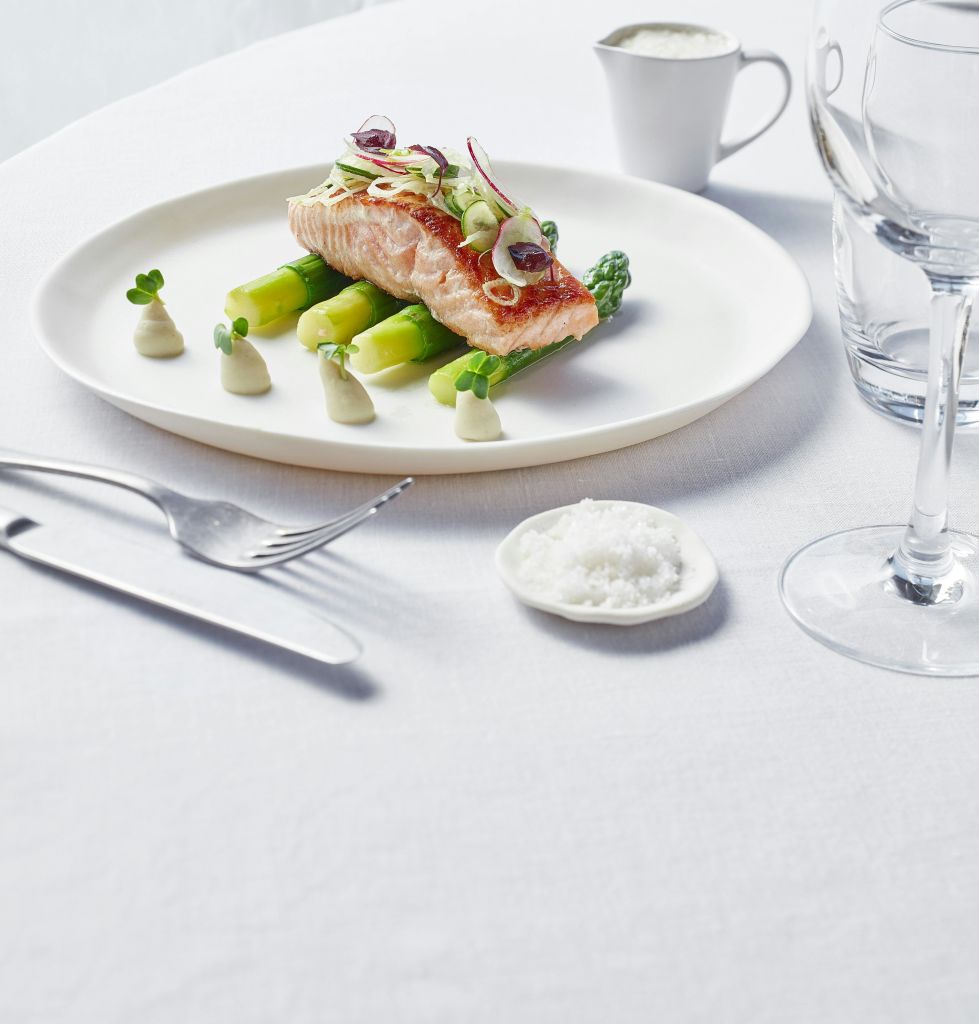 장식이 있는 아스파라거스의 연어 필레는 식기와 유리잔, 하얀 식탁보에 담긴 작은 소금 접시가 담긴 흰색 접시에 담겨 제공됩니다.