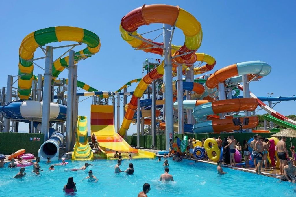 형형색색의 미끄럼틀이 있는 워터파크와 맑고 푸른 하늘 아래에서 물놀이를 즐기는 사람들로 가득한 수영장.