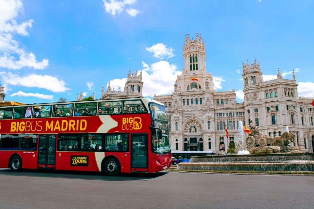 '마드리드 빅 버스' 라는 이름의 빨간색 2층 투어 버스가 푸른 하늘 아래 웅장하고 화려한 건물을 지나갑니다.
