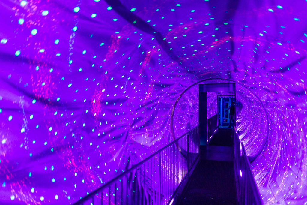 금속 난간이 있는 통로를 따라 선명한 보라색 조명과 청록색 점이 있는 터널.