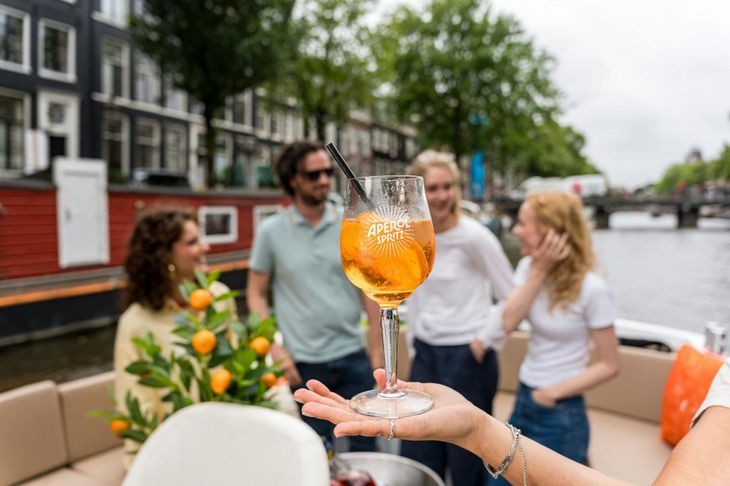 건물과 나무를 배경으로 보트에서 이야기하는 네 사람 앞에서 아페롤 스프리츠를 들고 있는 손.
