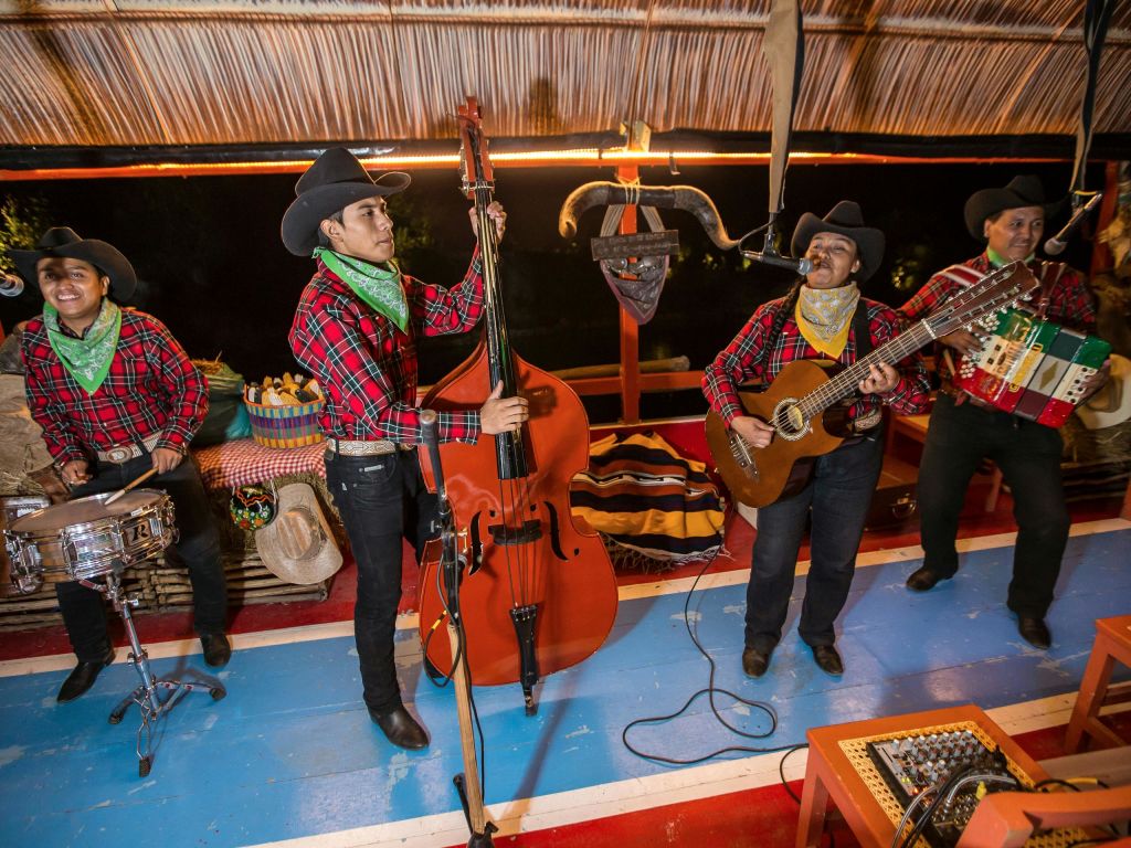 카우보이 모자와 격자무늬 셔츠를 입은 세 명의 뮤지션이 다채로운 무대에서 더블 베이스, 기타, 드럼을 들고 공연합니다.