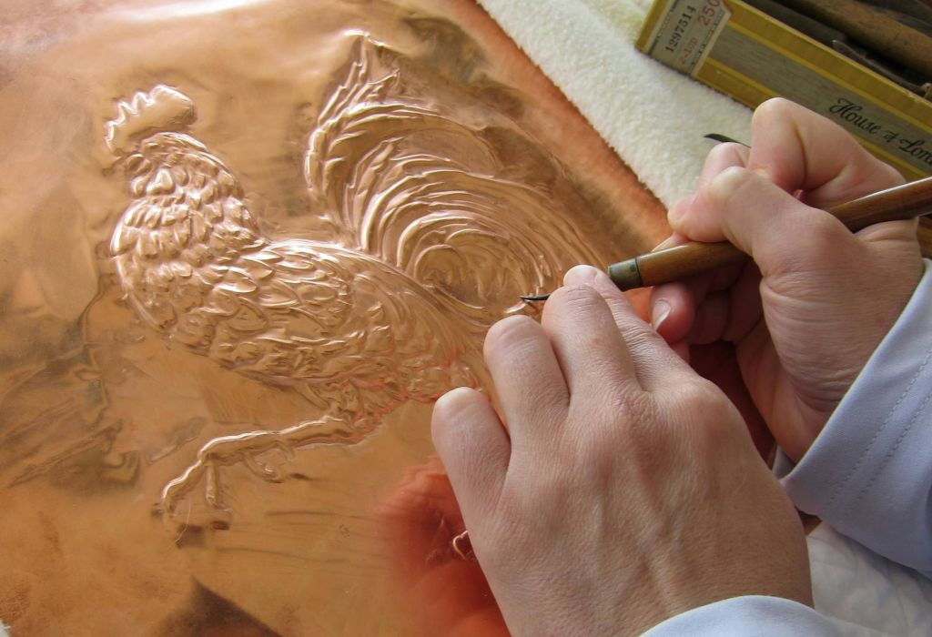 구리 시트에 수탉 디자인을 새기는 도구를 사용하는 손. 시트 옆에는 도구 상자가 있습니다.