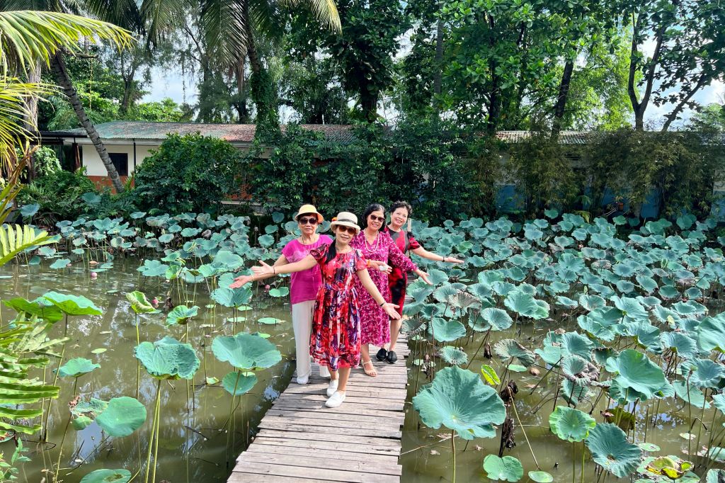 화사한 색상의 옷을 입은 네 명의 여성이 녹음으로 둘러싸인 백합 연못 위 나무 산책로에서 포즈를 취하고 있습니다.