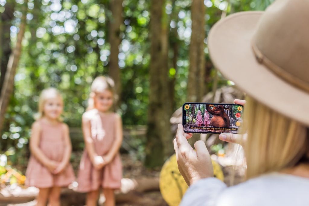분홍색 드레스를 입은 두 명의 어린 소녀가 숲 속에서 사진을 찍고 있습니다. 사진을 찍는 스마트폰에 중점을 둡니다.