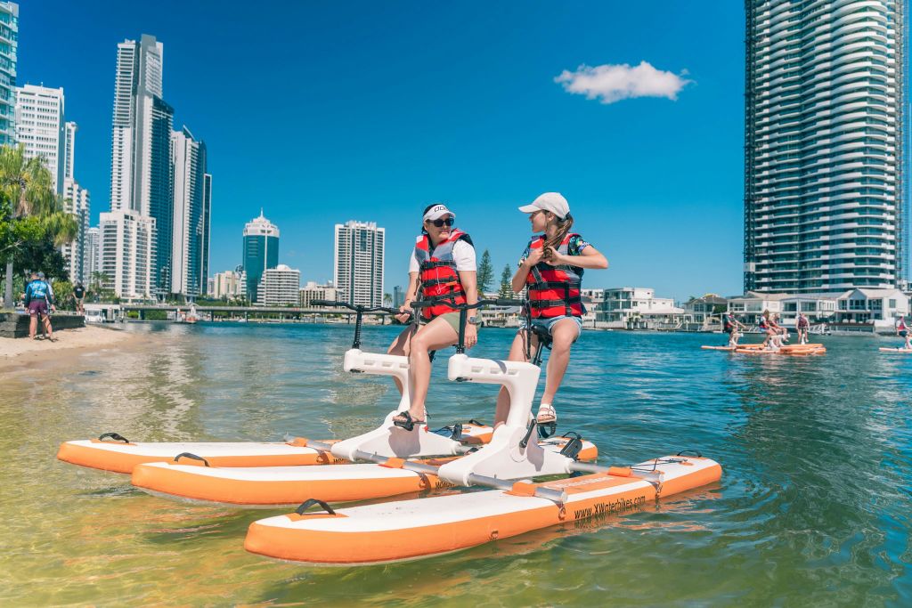 구명조끼를 입은 두 사람이 도시의 스카이라인을 배경으로 차분한 물가에서 수상 자전거를 타고 있습니다.