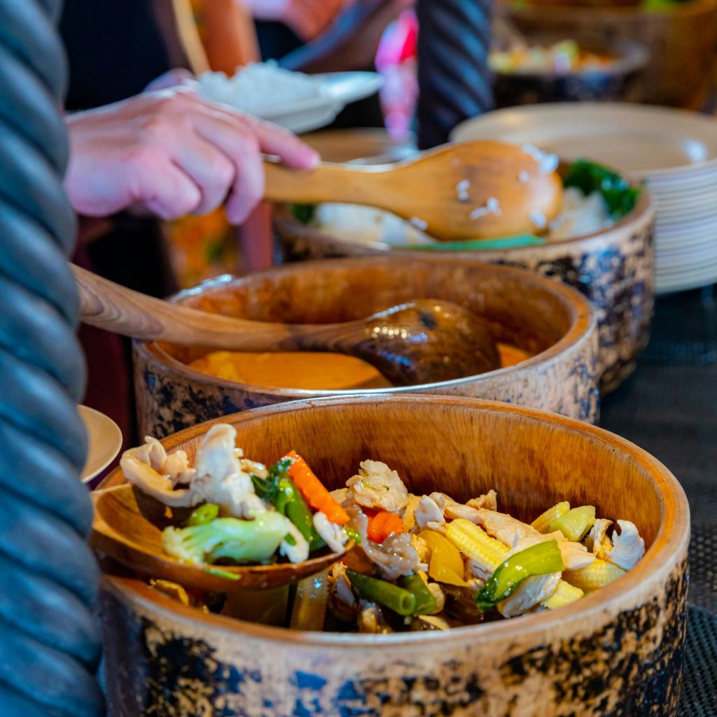 한 사람이 나무 국자를 사용하여 다양한 조리된 야채로 채워진 크고 소박한 나무 그릇의 음식을 제공합니다. 접시는 배경에 있습니다.