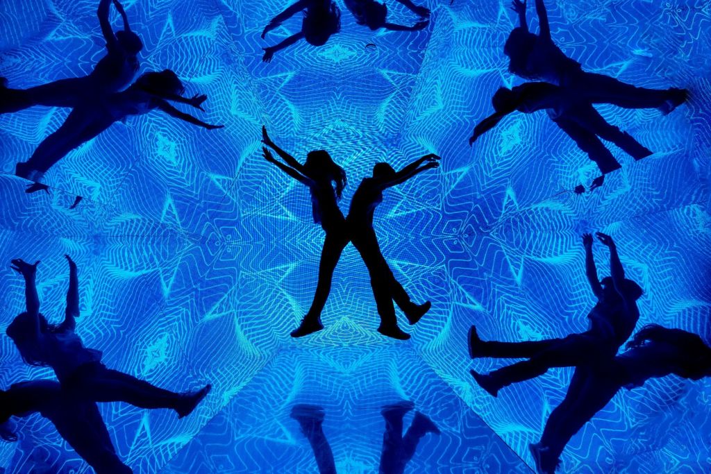추상적인 기하학적 패턴과 반사가 돋보이는 선명한 파란색 배경에 맞춰 춤추는 두 사람의 실루엣.