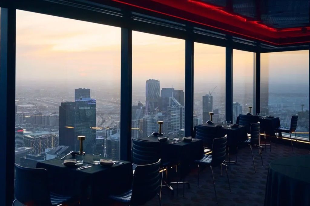 해질녘 도시의 스카이라인이 내려다보이는 바닥에서 천장까지 이어지는 창문이 있는 현대적인 레스토랑입니다.