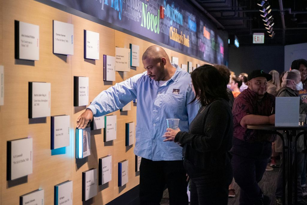 텍스트 디스플레이가 있는 벽을 바라보는 두 사람, 한 사람은 컵을 들고 있습니다. 설정이 전시회나 박물관인 것 같습니다.