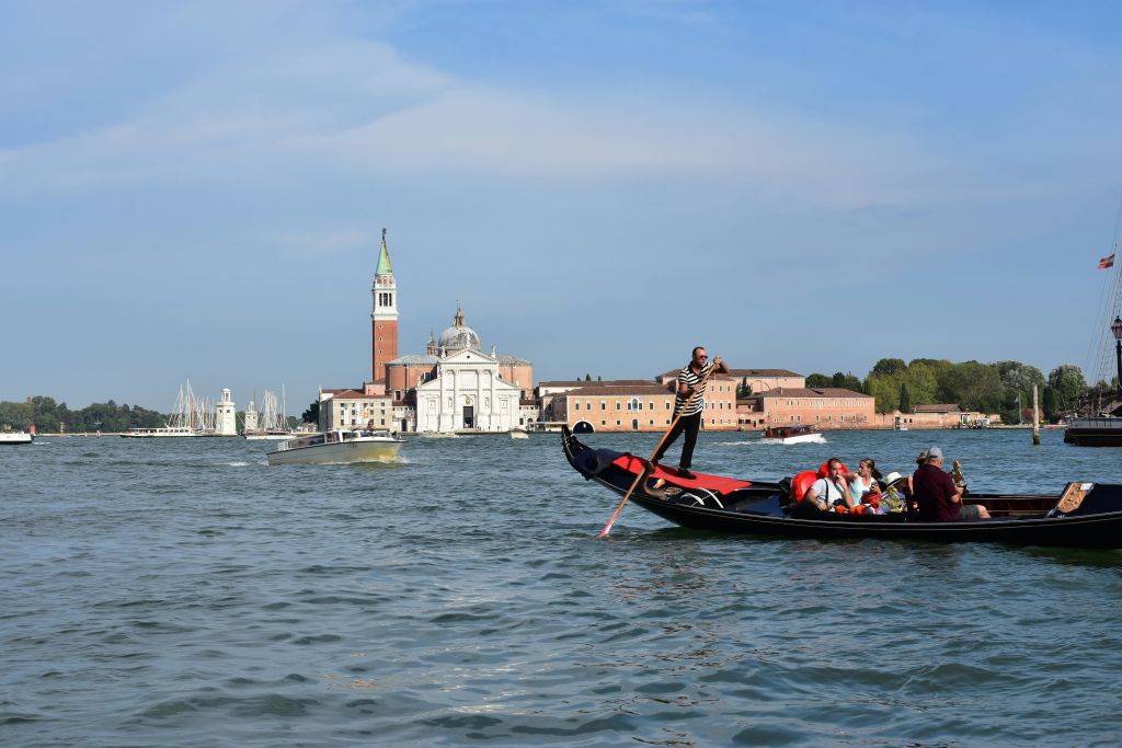 곤돌라이어는 높은 탑과 교회를 포함한 유서 깊은 건물 앞의 운하에서 승객과 함께 곤돌라를 젓고 있습니다.